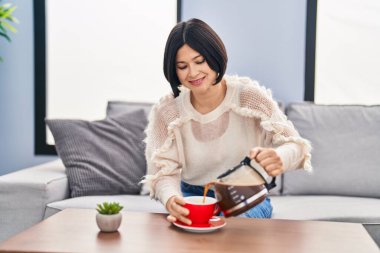 Young chinese woman pouring coffee on cup sitting on sofa at home