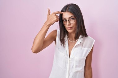 Brunette young woman standing over pink background wearing glasses pointing unhappy to pimple on forehead, ugly infection of blackhead. acne and skin problem 