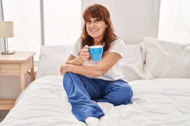 Orta yaşlı bir kadın yatak odasında bir fincan kahve içiyor.