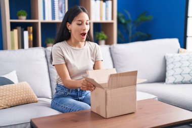 Young hispanic woman opening cardboard box afraid and shocked with surprise and amazed expression, fear and excited face. 