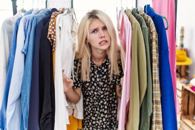 Young blonde woman searching clothes on clothing rack clueless and confused expression. doubt concept. 