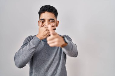 Hispanic man with beard standing over white background laughing at you, pointing finger to the camera with hand over mouth, shame expression 