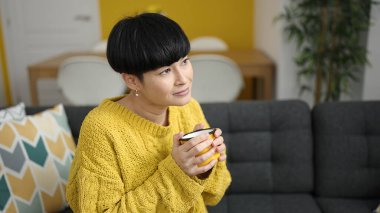 Genç Çinli kadın evdeki kanepede oturup kahve içiyor.