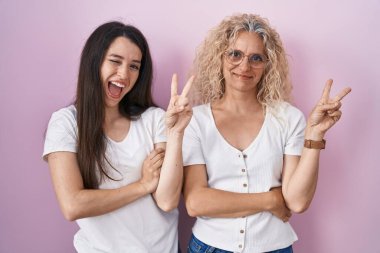 Anne ve kızı pembe arka planda yan yana durup gülümsüyor ve kameraya göz kırpıyor parmaklarıyla zafer işareti yapıyor. İki numara.. 