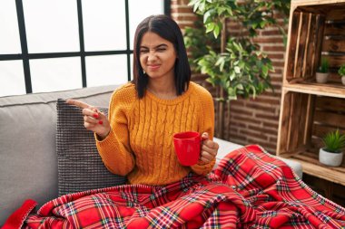 Young hispanic woman sitting on the sofa drinking a coffee at home pointing aside worried and nervous with forefinger, concerned and surprised expression 