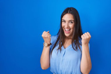 Mavi arka planda duran genç esmer kadın çok mutlu ve heyecanlı kollarını kaldırıp, gülümseyerek ve başarı için bağırarak kazanan jestler yapıyor. kutlama konsepti. 