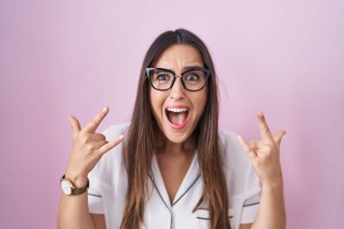 Young brunette woman wearing glasses standing over pink background shouting with crazy expression doing rock symbol with hands up. music star. heavy concept. 