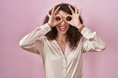 Hispanic woman with curly hair standing over pink background doing ok gesture like binoculars sticking tongue out, eyes looking through fingers. crazy expression. 