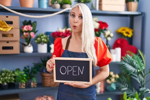 Caucasian woman working at florist holding open sign in shock face, looking skeptical and sarcastic, surprised with open mouth