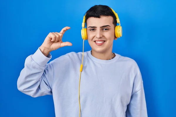 Non binary person listening to music using headphones smiling and confident gesturing with hand doing small size sign with fingers looking and the camera. measure concept.
