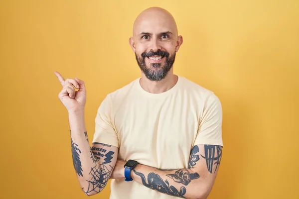 Hispanic man with tattoos standing over yellow background with a big smile on face, pointing with hand finger to the side looking at the camera.