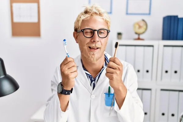 Young Blond Dentist Man Working Dentist Clinic Holding Toothbrushes Winking — Photo