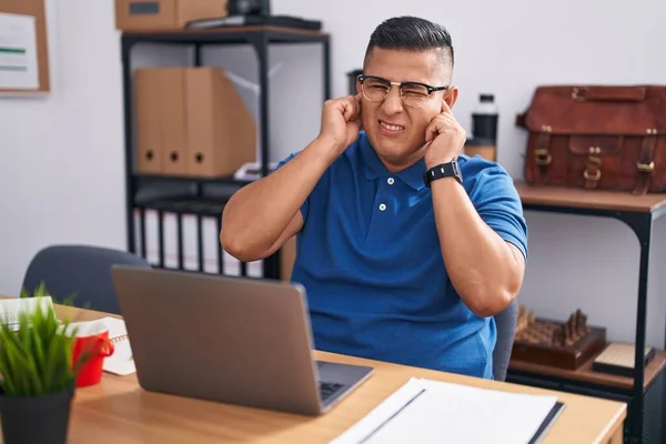 Young hispanic man working at the office with laptop covering ears with fingers with annoyed expression for the noise of loud music. deaf concept.
