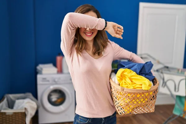 Young woman holding laundry basket smiling cheerful playing peek a boo with hands showing face. surprised and exited