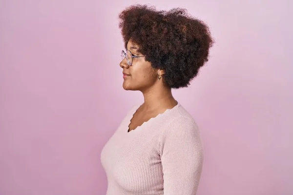 Young african american woman standing over pink background looking to side, relax profile pose with natural face and confident smile.