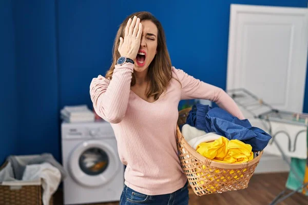 Young woman holding laundry basket yawning tired covering half face, eye and mouth with hand. face hurts in pain.