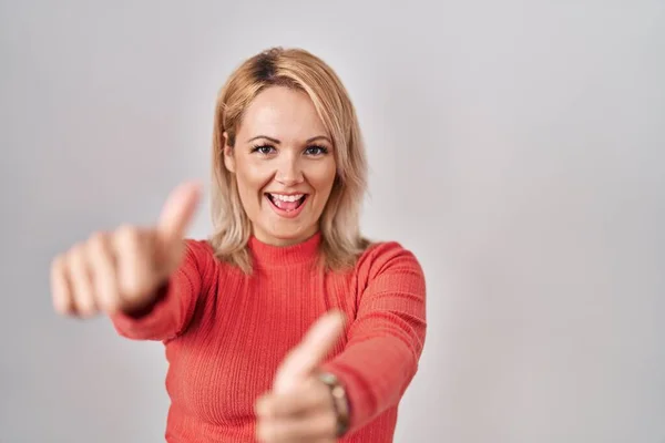 Blonde woman standing over isolated background approving doing positive gesture with hand, thumbs up smiling and happy for success. winner gesture.