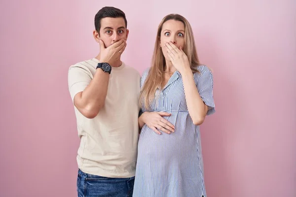 Young Couple Expecting Baby Standing Pink Background Shocked Covering Mouth — Stock Photo, Image