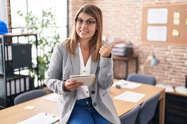 Young hispanic woman working at the office wearing glasses pointing to the back behind with hand and thumbs up, smiling confident