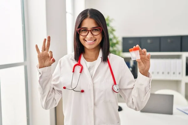 Young Hispanic Doctor Woman Holding Sample Cup Doing Sign Fingers — ストック写真