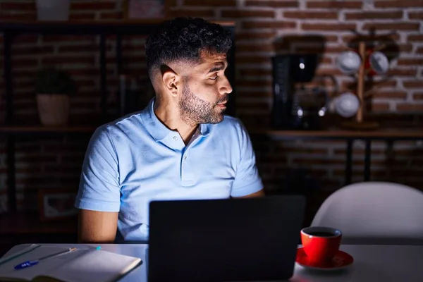 Hispanic man with beard using laptop at night looking to side, relax profile pose with natural face with confident smile.