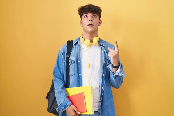 Hispanic teenager wearing student backpack and holding books amazed and surprised looking up and pointing with fingers and raised arms.