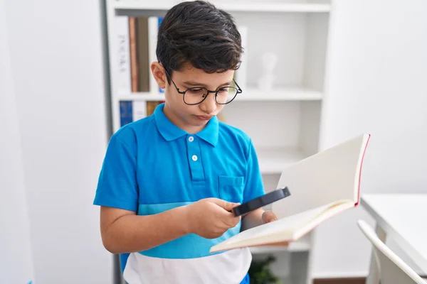 Adorable hispanic boy student reading book using magnifying glass at library school