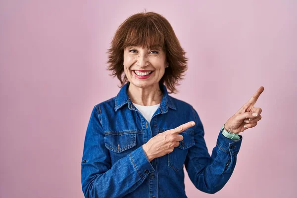 Middle age woman standing over pink background smiling and looking at the camera pointing with two hands and fingers to the side.