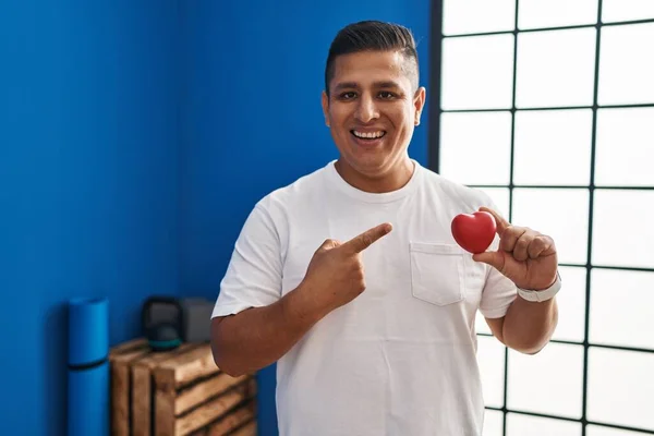 Hispanic young man holding red heart at gym smiling happy pointing with hand and finger