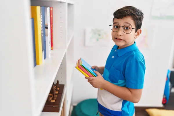 Adorable hispanic boy student smiling confident holding books of shelving at library school