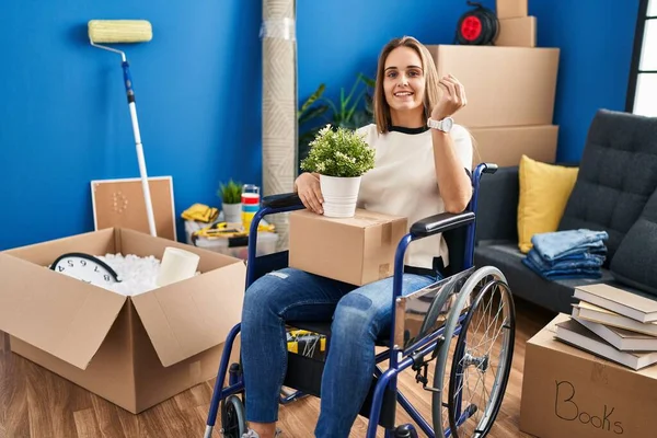 Young woman sitting on wheelchair moving to a new home doing money gesture with hands, asking for salary payment, millionaire business