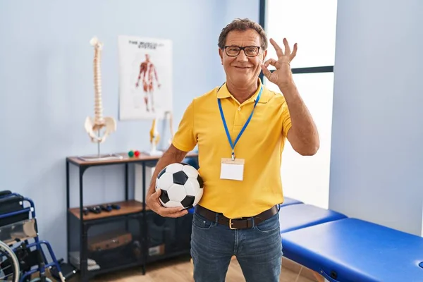 Senior man working at sport physiotherapy clinic doing ok sign with fingers, smiling friendly gesturing excellent symbol