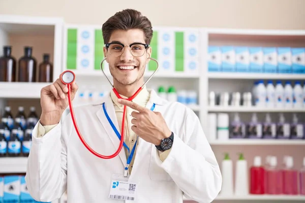 Young hispanic man working at pharmacy drugstore using stethoscope smiling happy pointing with hand and finger