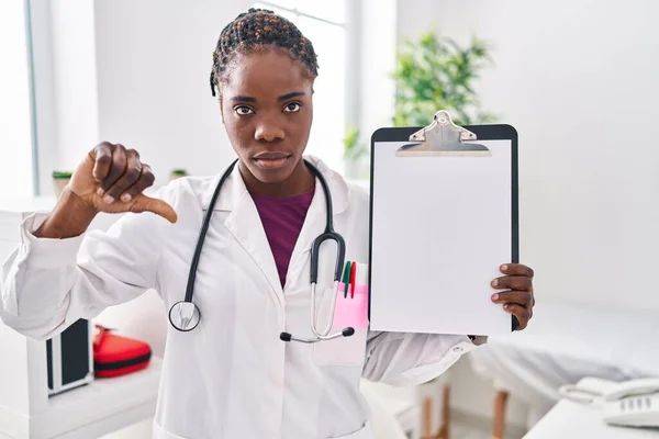 Beautiful black doctor woman holding clipboard with angry face, negative sign showing dislike with thumbs down, rejection concept