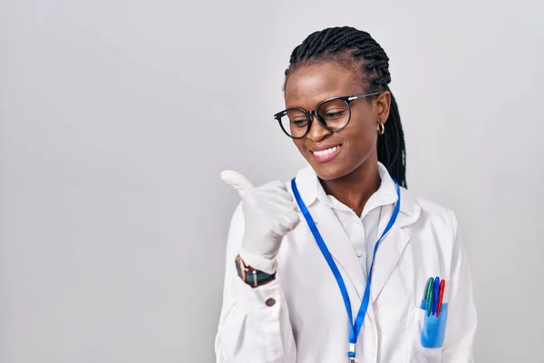 African Woman Braids Wearing Scientist Uniform Pointing Thumb Side Smiling — Stockfoto