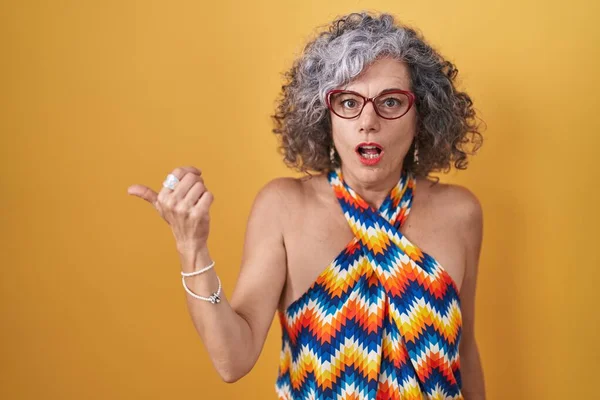 Middle age woman with grey hair standing over yellow background surprised pointing with hand finger to the side, open mouth amazed expression.