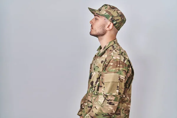 Young hispanic man wearing camouflage army uniform looking to side, relax profile pose with natural face with confident smile.