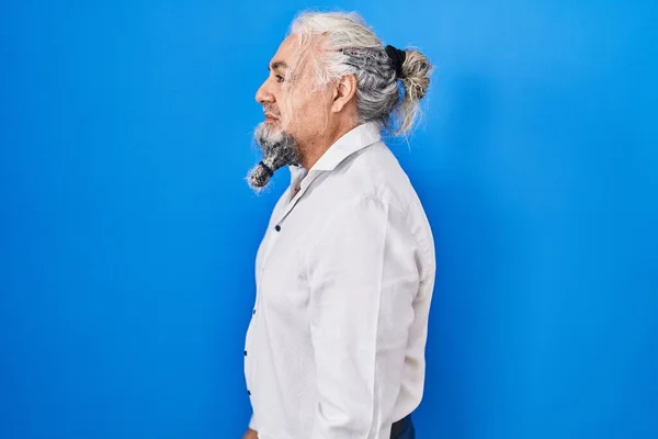 Middle age man with grey hair standing over blue background looking to side, relax profile pose with natural face and confident smile.