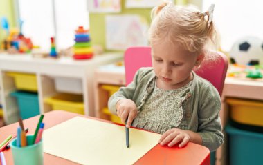 Adorable blonde girl preschool student sitting on table drawing on paper at kindergarten
