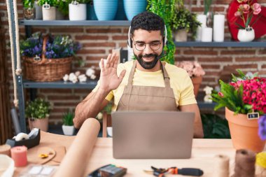 Hispanic man with beard working at florist shop doing video call looking positive and happy standing and smiling with a confident smile showing teeth 