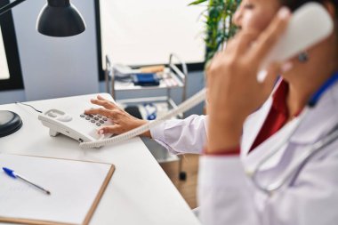 Orta yaşlı İspanyol kadın. Doktor üniforması giyiyor. Klinikte telefonla konuşuyor.