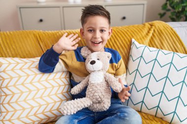 Adorable hispanic boy saying hello with hand sitting on sofa at home