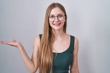 Young caucasian woman standing over white background smiling cheerful presenting and pointing with palm of hand looking at the camera. 