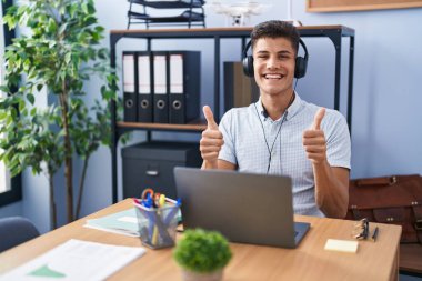 Young hispanic man working at the office wearing headphones success sign doing positive gesture with hand, thumbs up smiling and happy. cheerful expression and winner gesture. 