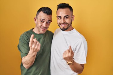 Homosexual couple standing over yellow background beckoning come here gesture with hand inviting welcoming happy and smiling 