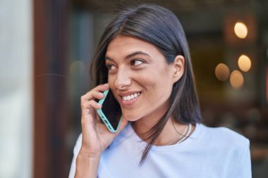 Genç, güzel, İspanyol bir kadın gülümsüyor ve caddede akıllı telefondan konuşuyor.