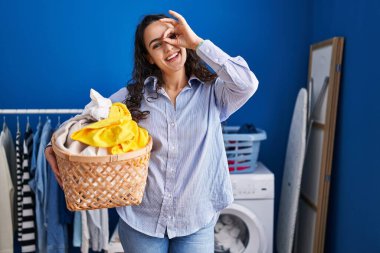 Young brunette woman holding laundry basket smiling happy doing ok sign with hand on eye looking through fingers 