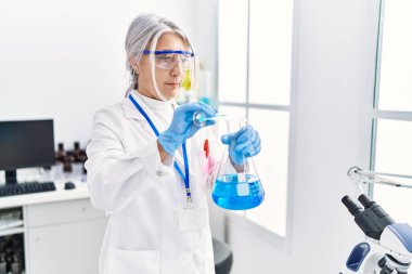 Orta yaşlı, gri saçlı, bilim adamı üniforması giyen bir kadın laboratuarda test tüpüne sıvı döküyor.