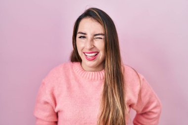 Young hispanic woman standing over pink background winking looking at the camera with sexy expression, cheerful and happy face. 