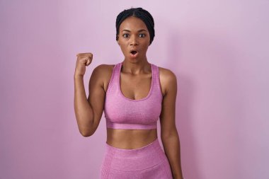 African american woman with braids wearing sportswear over pink background surprised pointing with hand finger to the side, open mouth amazed expression. 